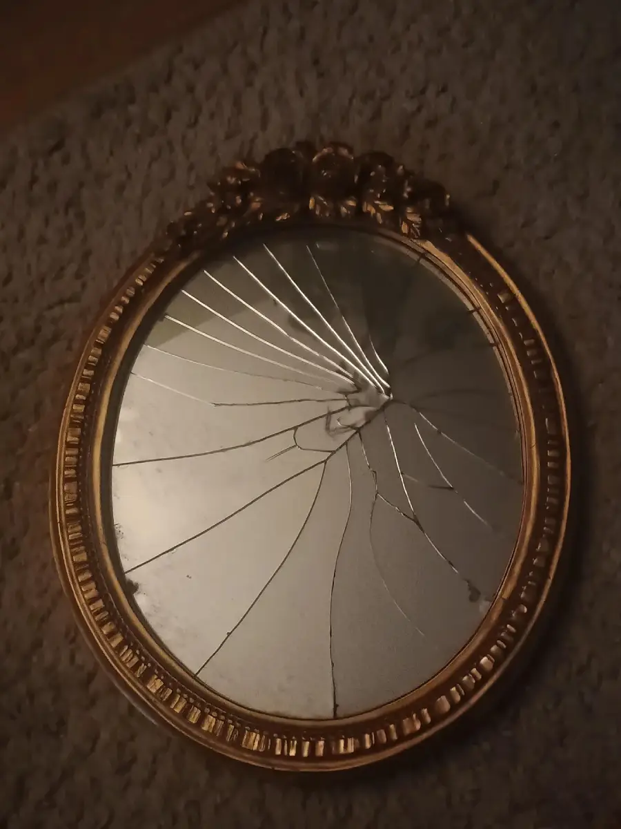 An oval, gold-framed mirror with ornate floral detailing at the top is lying on a carpeted floor. The mirror glass is shattered with cracks radiating from the center.