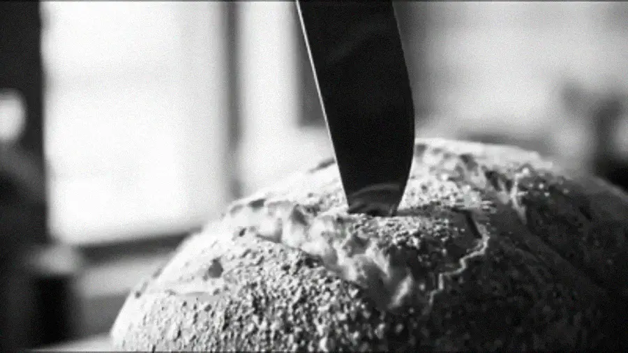 A close-up, black-and-white image of a serrated knife slicing into a round loaf of rustic bread with a textured crust.