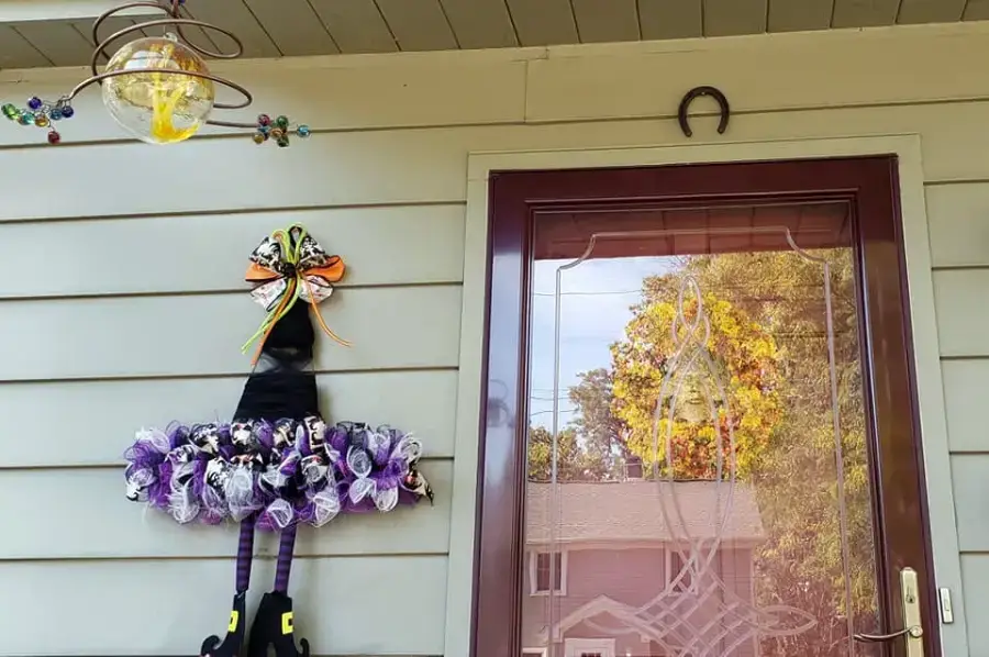 A whimsical Halloween decoration shaped like a witch’s hat, skirt, and legs hangs on a house’s exterior wall beside a glass door. Above is a horseshoe and a glass ornament. Trees and houses are reflected in the door.