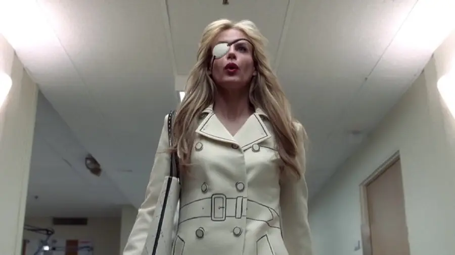 A woman with long blonde hair, wearing an off-white trench coat and an eye patch, walks confidently down a brightly lit hallway.