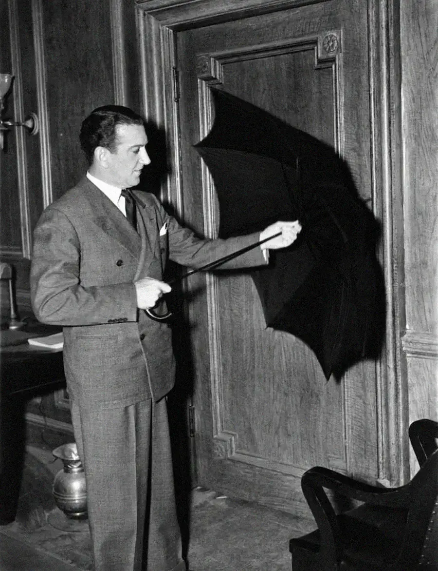 A man in a suit stands indoors, holding an open umbrella and pointing it toward a closed wooden door, as if demonstrating or inspecting it.