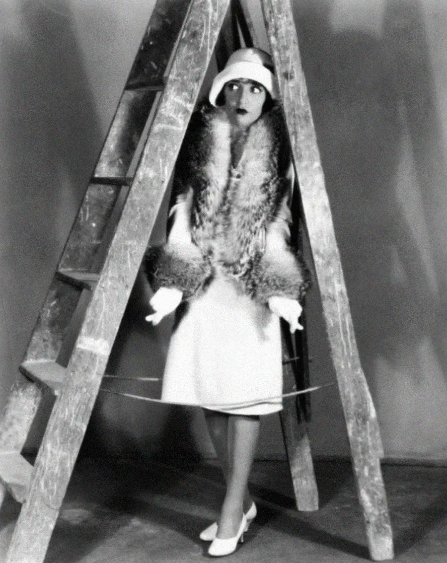 A woman in 1920s attire, including a cloche hat, fur stole, and white dress, stands between a wooden ladder, looking to the side with a surprised expression. The photo is black and white.