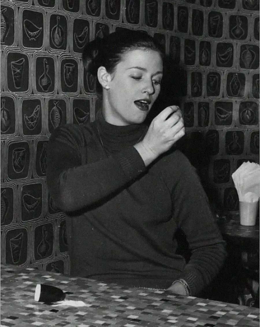 A woman with dark hair in a bun, wearing a long-sleeve turtleneck, sits at a tiled table in front of a patterned wall, holding her hand up and looking at it with her mouth slightly open.