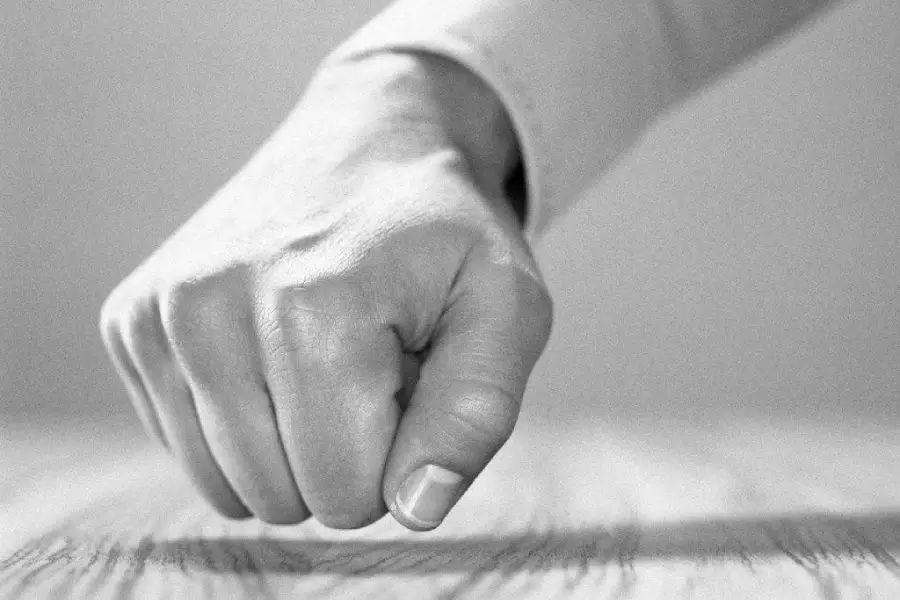 A close-up of a person's hand in a fist, resting or pounding on a wooden surface. The image is in black and white.
