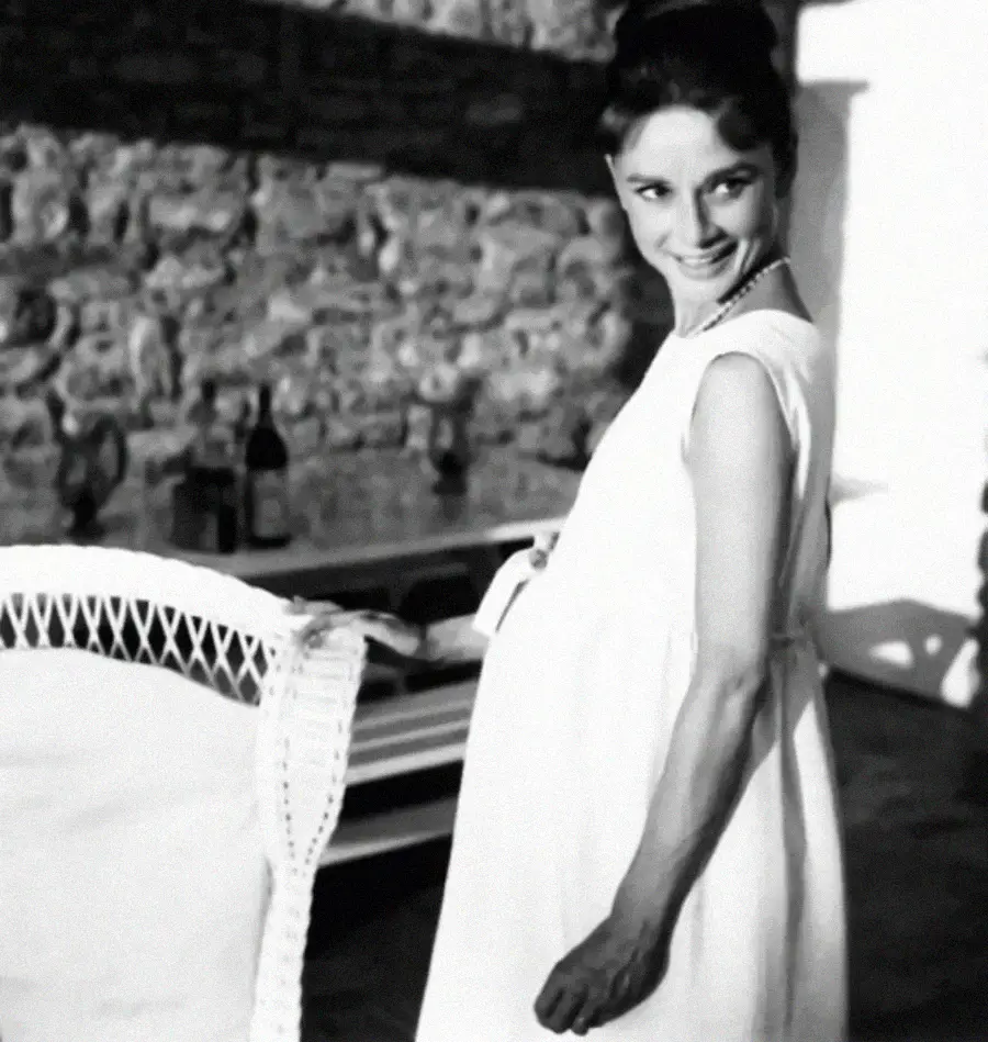 A woman in a sleeveless white dress stands and smiles, holding her stomach. Behind her is a stone wall, a table, and some bottles. The setting appears bright and relaxed.