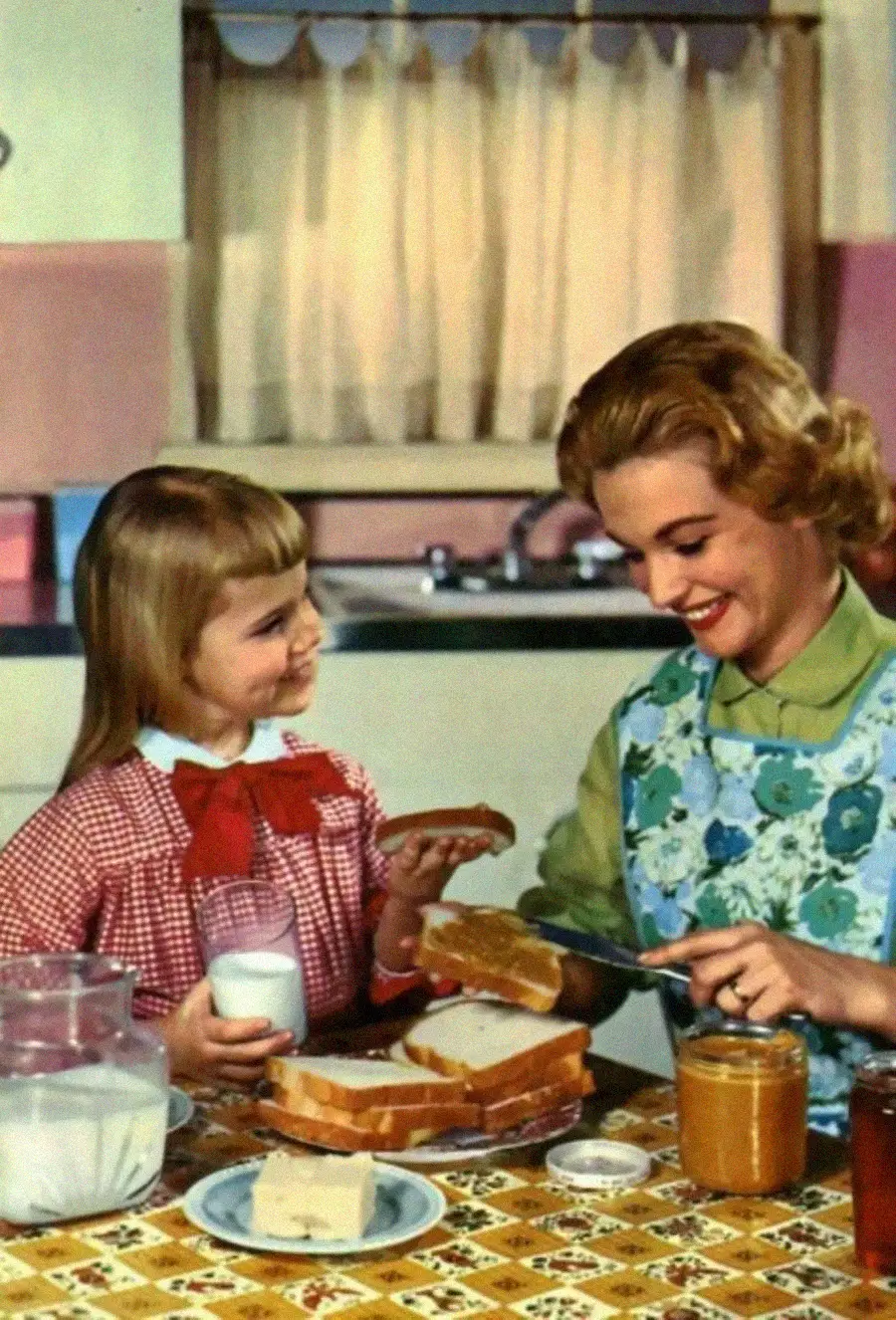 A woman in an apron spreads peanut butter on bread while smiling at a young girl holding a glass of milk. They are in a vintage kitchen with pink walls and patterned tablecloth.