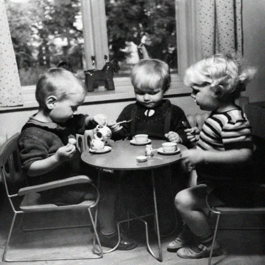Three young children sit around a small table having a tea party with a toy tea set. They appear focused on pouring tea and holding cups. The scene is indoors near a window with curtains.