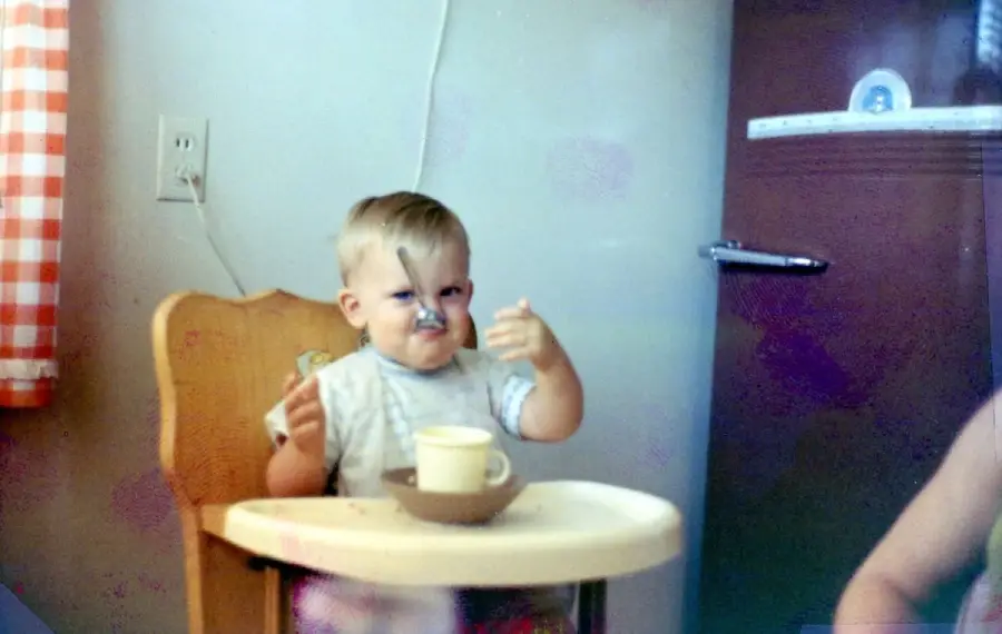A toddler sits in a high chair, holding a spoon to their nose with a cup and bowl on the tray. The kitchen features a door, power outlet, and a window with red-and-white checkered curtains.