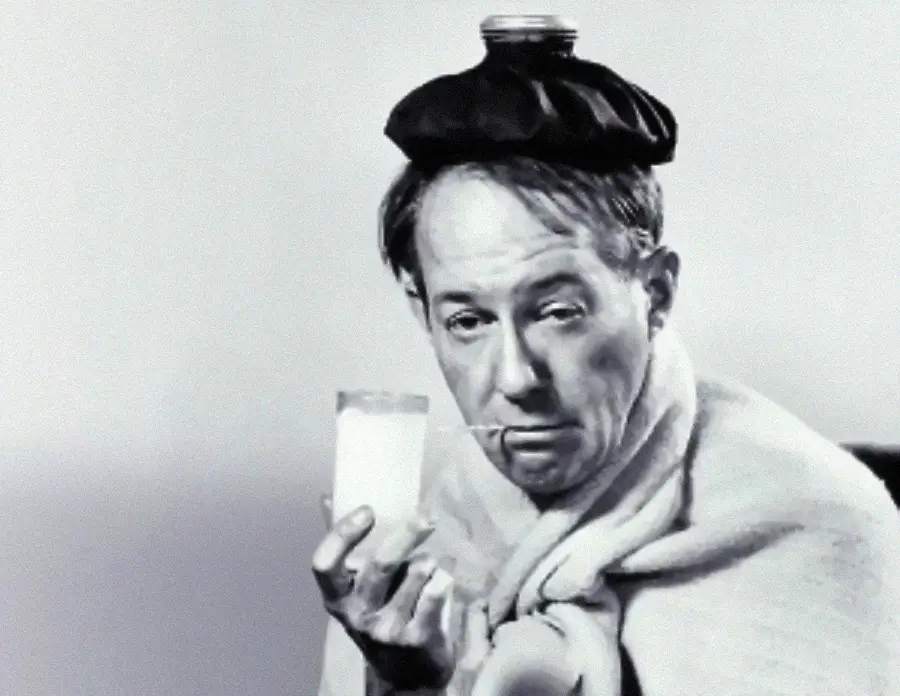 A sick person with a blanket, ice pack on their head, holding a glass with a straw, and looking tired.