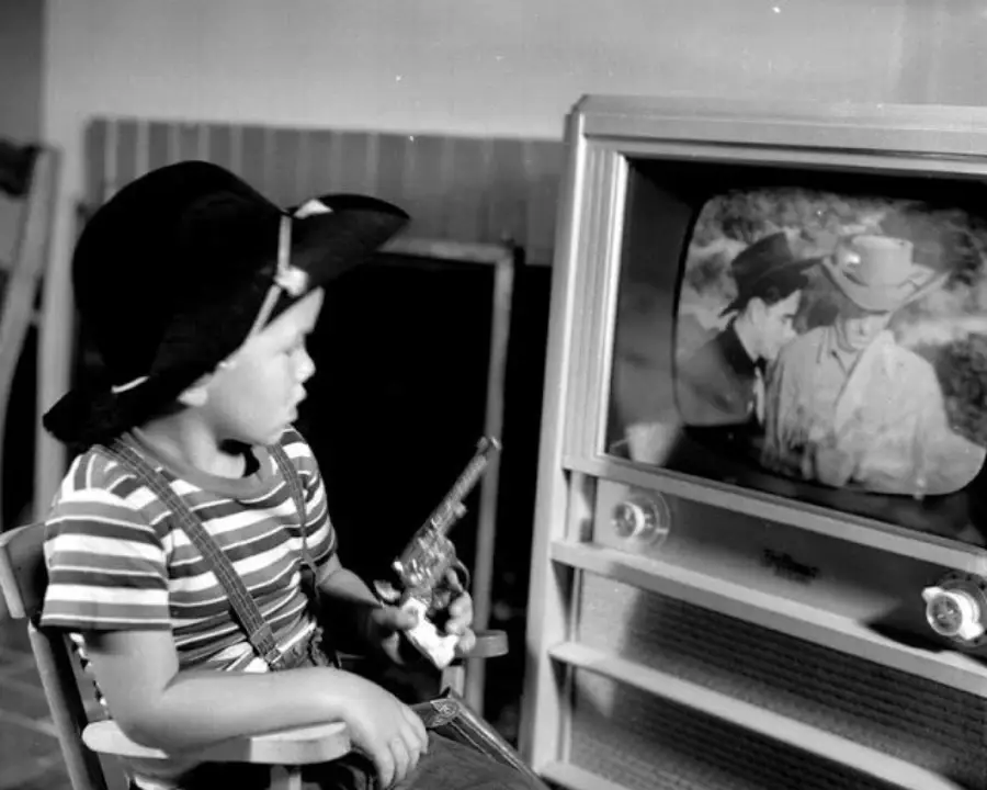 A young child in a cowboy hat and striped shirt watches a black-and-white TV show featuring cowboys, holding a toy gun and sitting in a small chair.
