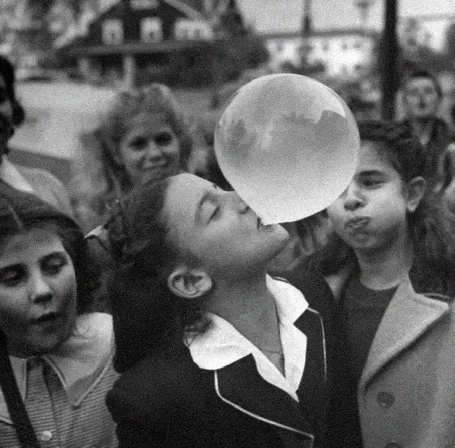 A girl blows a large bubblegum bubble while friends watch and smile outdoors, with houses and trees in the background. The image is black and white, creating a nostalgic, vintage feel.