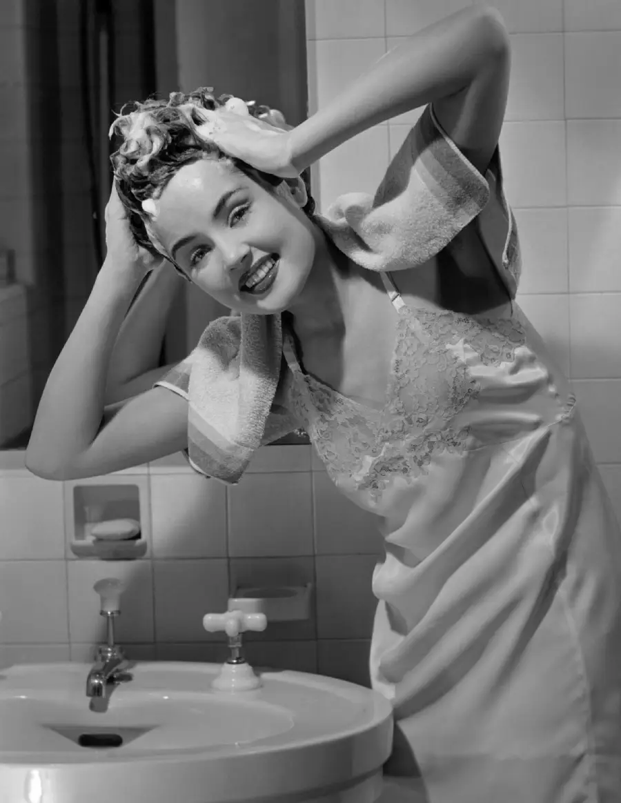 Smiling woman in a nightgown shampoos her hair over a bathroom sink, with a towel draped over her shoulders in a black-and-white photo.