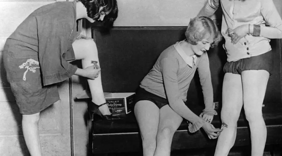 Three women in vintage clothing shave their legs together; one is standing with her leg up, while the other two sit on a bench, shaving. A can labeled "Valet Auto Strop" is visible on the bench. The image is black and white.