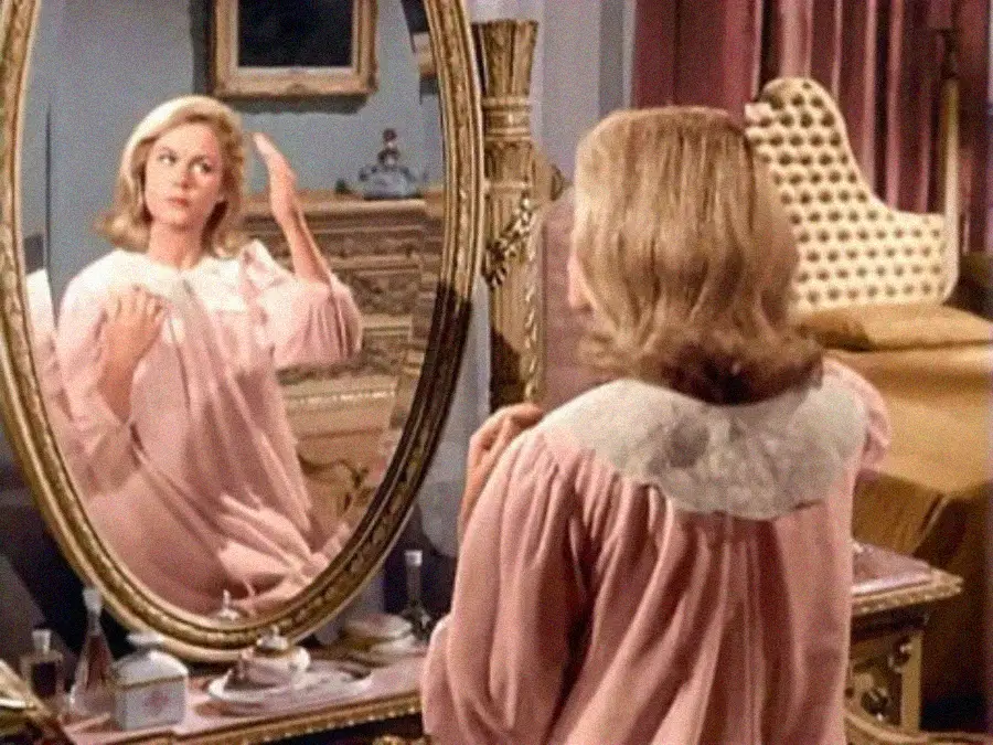 A woman in a pink nightgown sits at a vanity table, brushing her blonde hair and looking into an ornate mirror in a vintage-style bedroom.