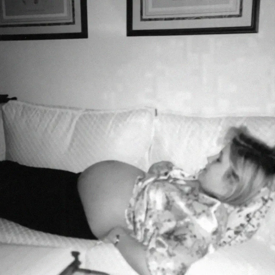 A pregnant person is lying on a couch, resting on their back with hands on their belly. The image is black and white, with framed pictures visible on the wall behind them.