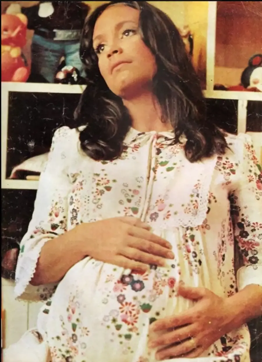 A pregnant woman with long dark hair wearing a floral dress rests her hands on her belly and looks thoughtful. She stands in front of shelves with toys and stuffed animals.