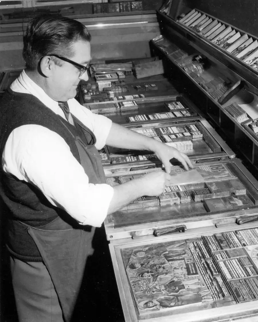 A man wearing glasses arranges metal type on a printing press, preparing materials for printing. He works carefully, surrounded by trays of type and printed images.