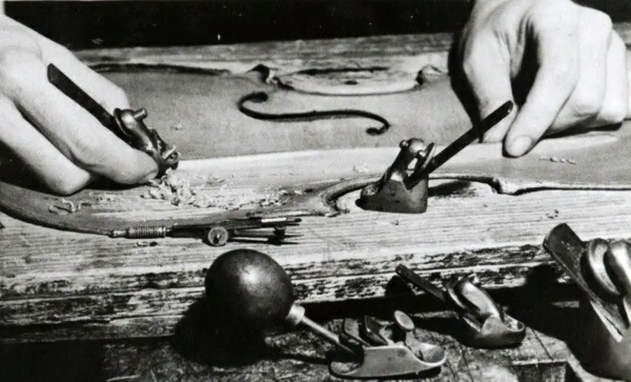 A person uses hand tools to carve the body of a violin from wood. Several small woodworking tools and shavings are visible on the workbench.