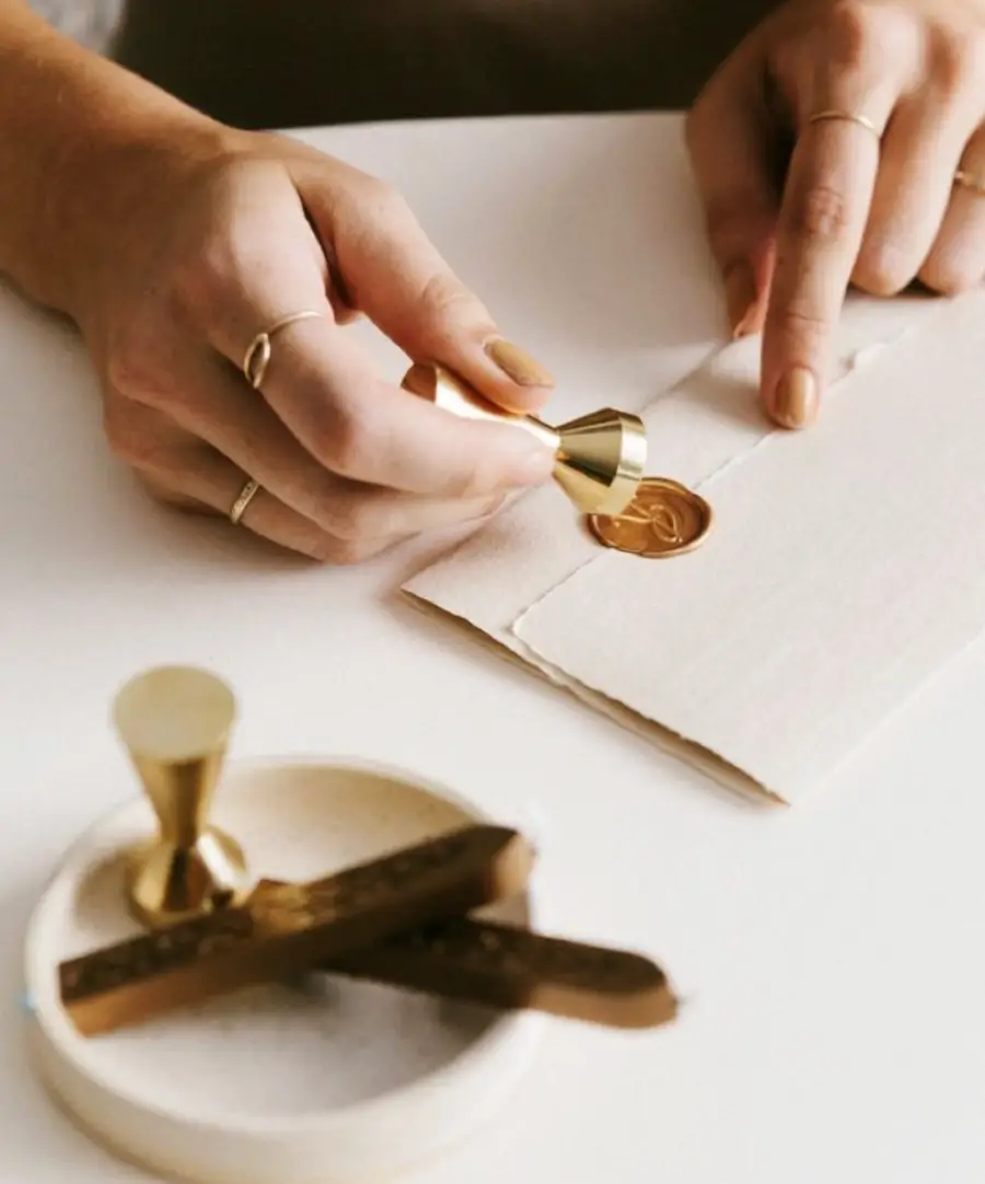 A person seals a cream-colored envelope with gold wax using a stamp; wax sticks and a seal stamp rest on a white dish nearby.