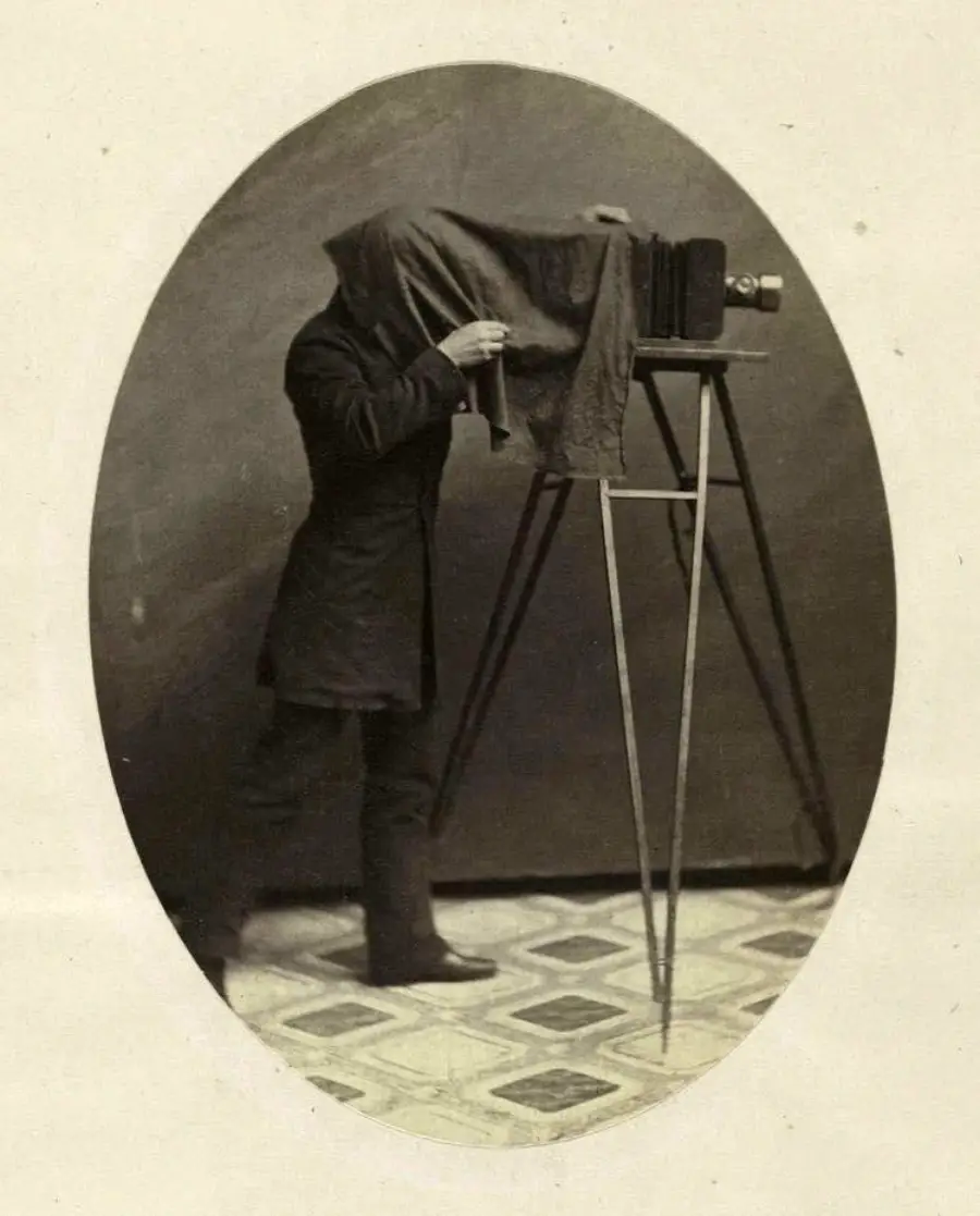 A person stands behind an old-fashioned camera on a tripod, covered by a dark cloth, preparing to take a photograph in a vintage studio setting.
