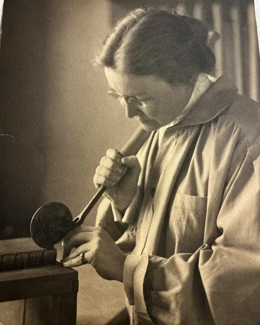 A person wearing glasses and a long coat carefully presses a tool onto the spine of a book, possibly performing bookbinding or restoration work.