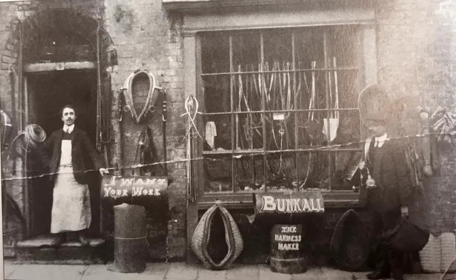 Black-and-white photo of two men outside a harness maker’s shop. Signs read "I WANT YOUR WORK" and "BUNKALL ZEEK HARNESS MAKER." Various harnesses and ropes hang on the shopfront and in the windows.