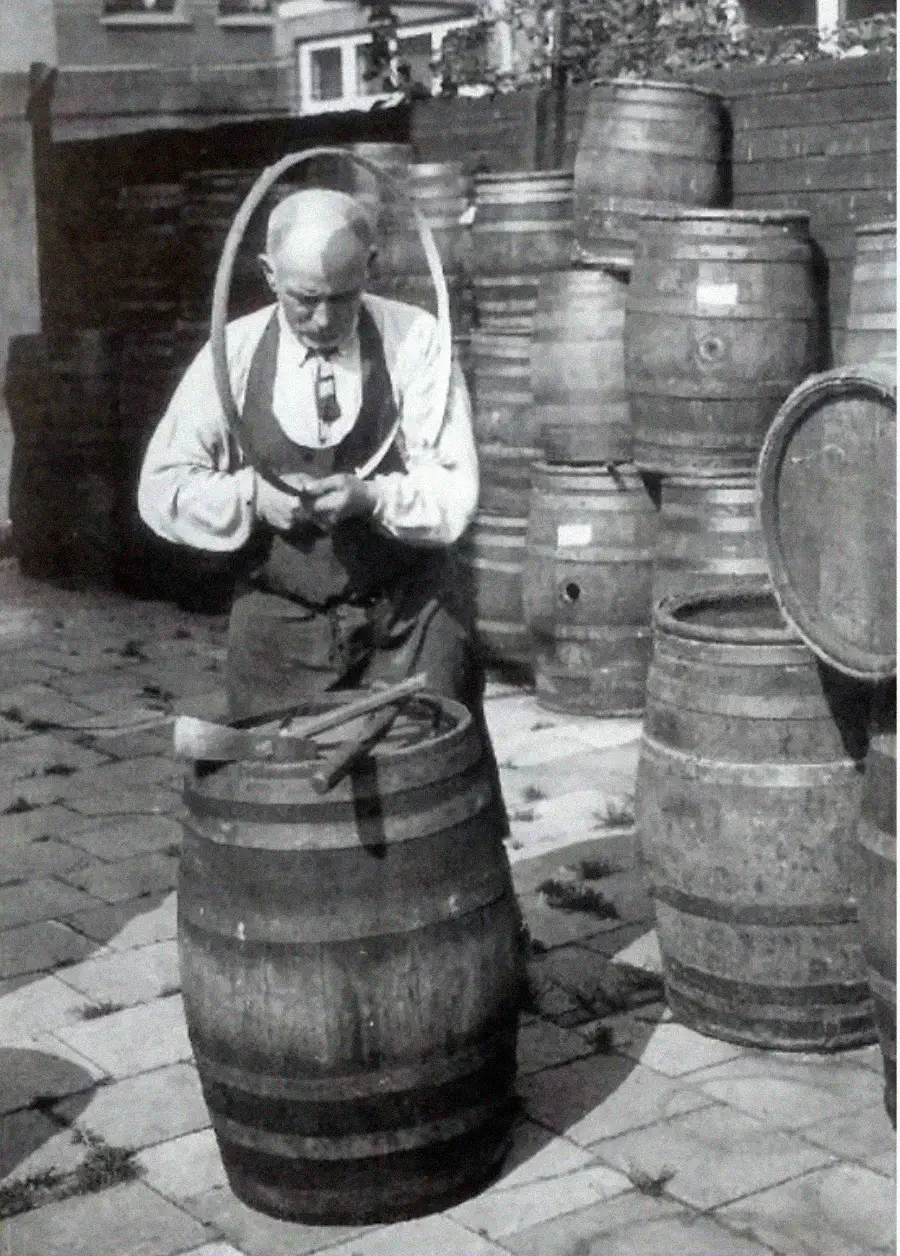 An older man works on a wooden barrel outdoors, holding a metal hoop. Several barrels and tools are around him, and a fence and houses are visible in the background.