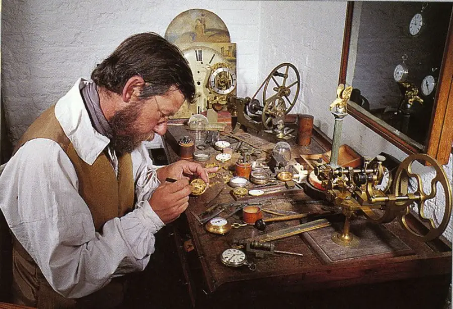 A man with dark hair and a beard repairs a pocket watch at a cluttered workbench filled with watch parts, tools, and vintage clock components.