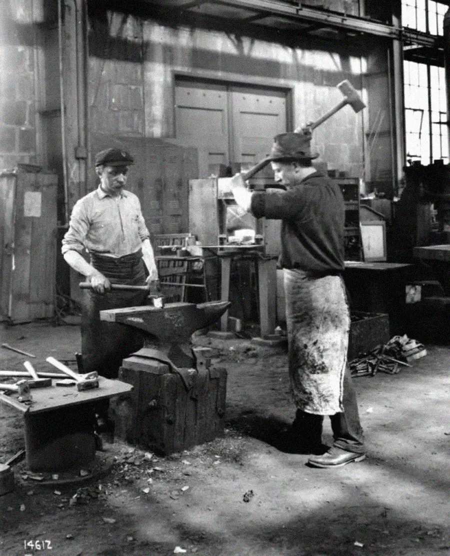 Two men working in a blacksmith shop: one holds metal with tongs on an anvil while the other, wearing an apron, raises a large hammer to strike. Tools and machinery fill the industrial background.