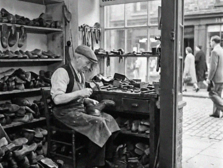 An elderly shoemaker wearing a cap and apron works on a shoe at a cluttered workbench in his shop, surrounded by shelves filled with various shoes. A window reveals people walking outside on the street.
