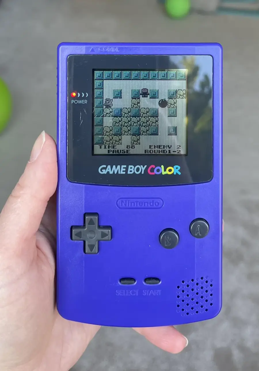 A hand holds a purple Game Boy Color displaying a pixelated game screen with blocks and characters. The device has black buttons and the Nintendo logo is visible below the screen.