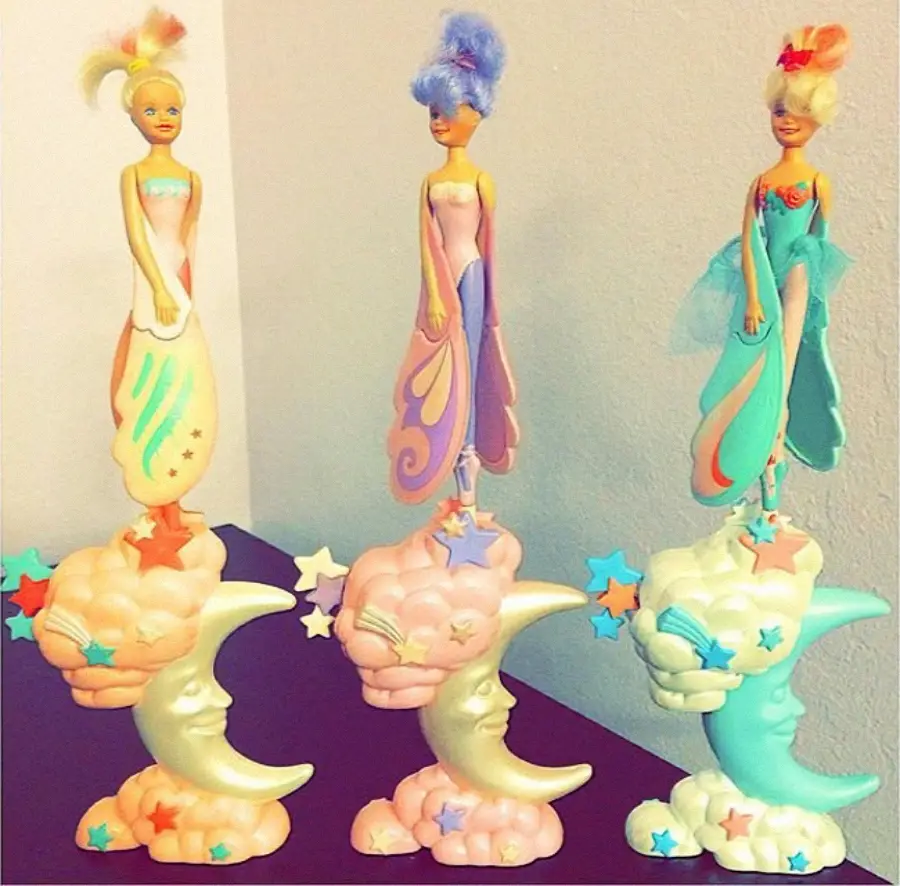 Three colorful dolls in pastel dresses stand atop crescent moon and cloud bases decorated with stars. Each doll has a distinct hairstyle and outfit, with whimsical, fantasy-themed designs. The scene is set against a plain wall.