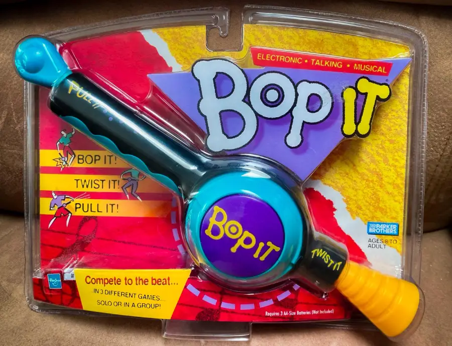 A packaged Bop It electronic game featuring a blue, yellow, and purple toy with levers labeled "Pull It!" and "Twist It!" The packaging promotes its musical, talking features and competitive gameplay.