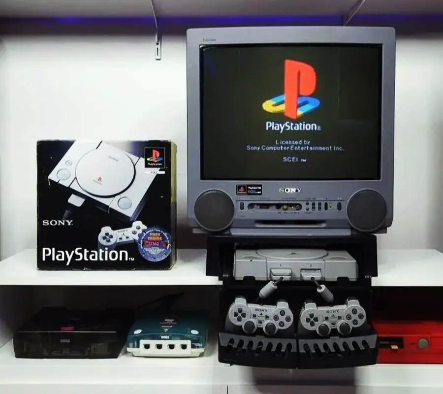 A retro PlayStation console setup with a CRT TV displaying the PlayStation logo. The PlayStation console sits below the TV with two controllers, and its original box is on the left. Other classic consoles are also visible on the shelf.