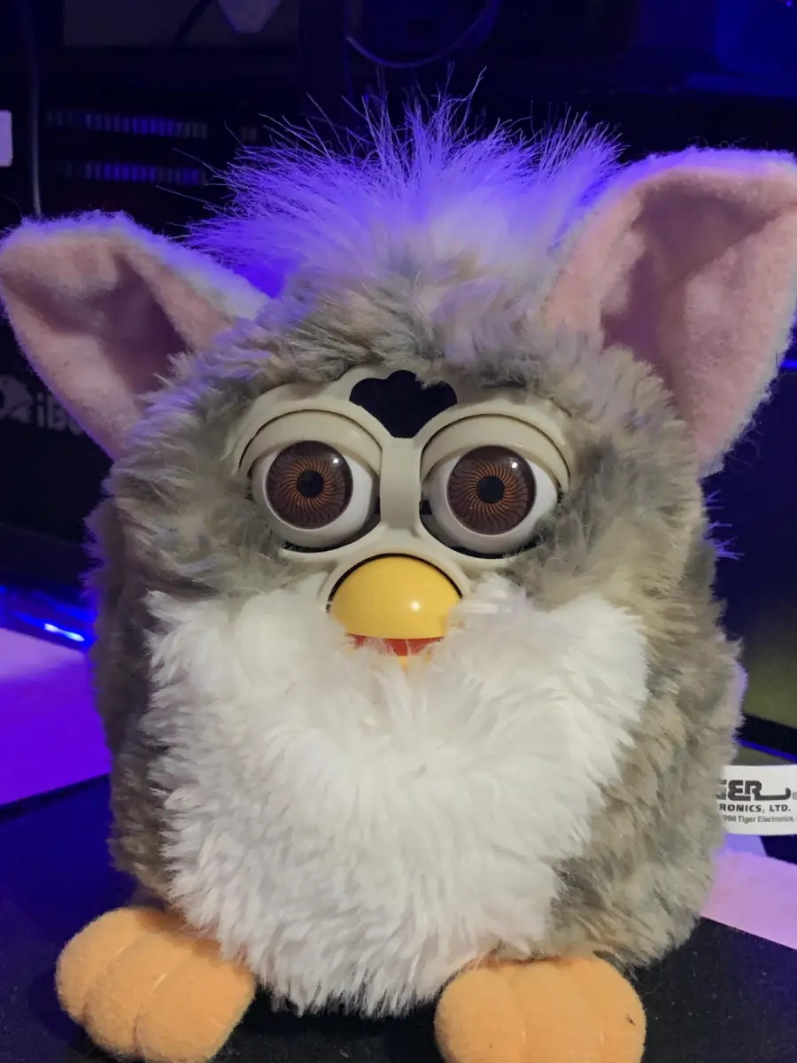 A close-up of a gray and white Furby toy with large, round eyes, yellow beak, pink ears, and orange feet, sitting on a surface with a purple-blue background.