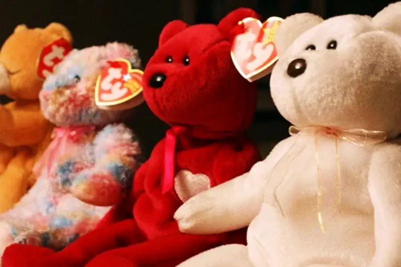 Four Beanie Baby teddy bears—yellow, multicolored, red, and white—sit in a row, each with a TY tag on its ear. The red bear has a heart on its chest. The background is dark, highlighting the bears.
