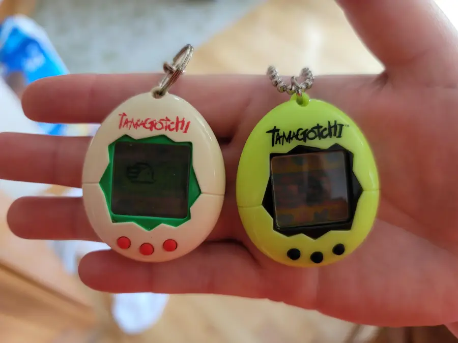 A hand holds two Tamagotchi digital pets, one white with green trim and the other bright green. Both have small screens and buttons, attached to keychains.