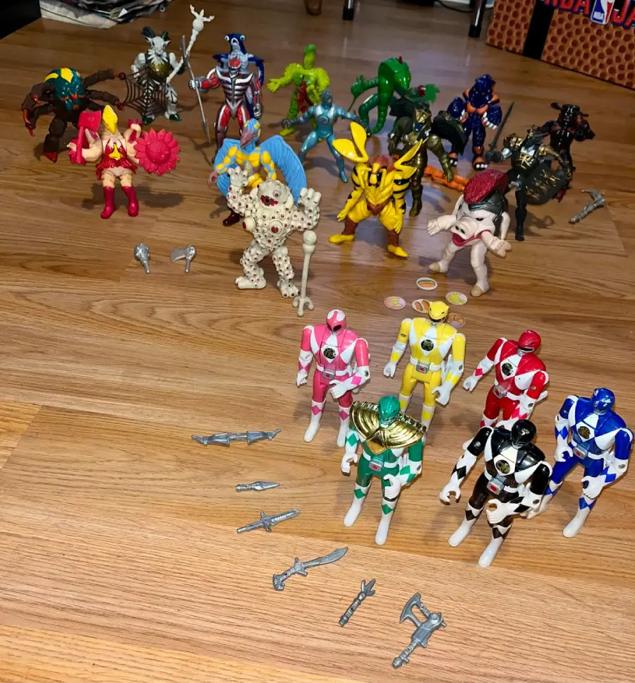 Several Power Rangers action figures and villain figures are arranged on a wooden floor, with various small weapons and accessories placed in front of them. The Power Rangers are in the front row.