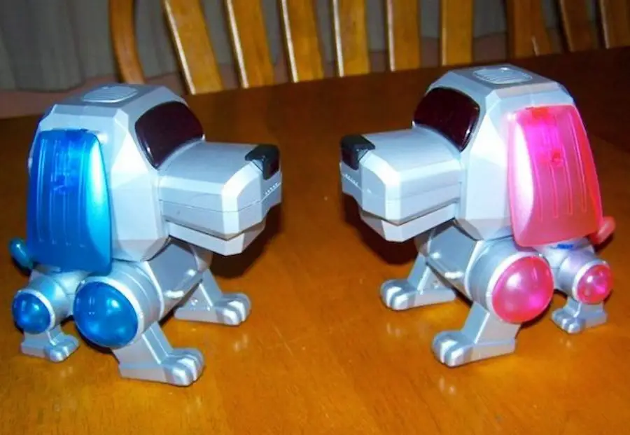 Two toy robot dogs sit facing each other on a wooden table. One has blue accents and the other has pink accents, with lights illuminated on their ears and hind legs. Wooden chairs are visible in the background.
