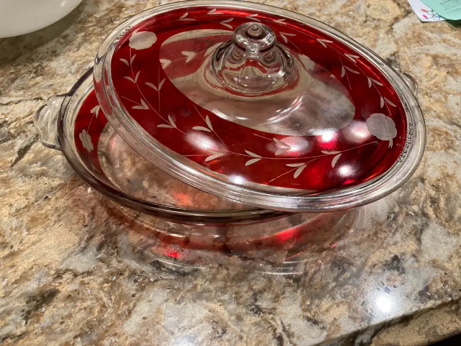 A glass casserole dish with a red and clear lid featuring etched leaf designs sits on a marbled countertop. The lid is partially open, revealing the empty interior of the dish.