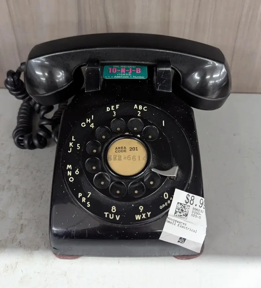 A black rotary dial telephone with a coiled cord sits on a gray surface. A price tag of $8.99 is attached to the phone, and the dial includes numbers and letters. The phone appears vintage.