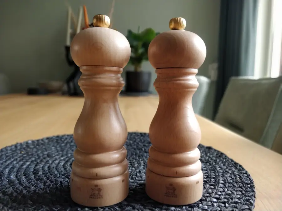 Two wooden pepper mills with gold knobs are standing side by side on a dark woven placemat atop a wooden dining table. The background shows blurred chairs, a plant, and soft natural light.