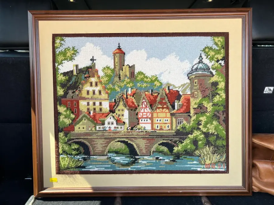 Framed needlepoint artwork of a European village, showing colorful buildings, a bridge over a river, trees, and a castle on a hill in the background. The scene is bordered with a light mat.
