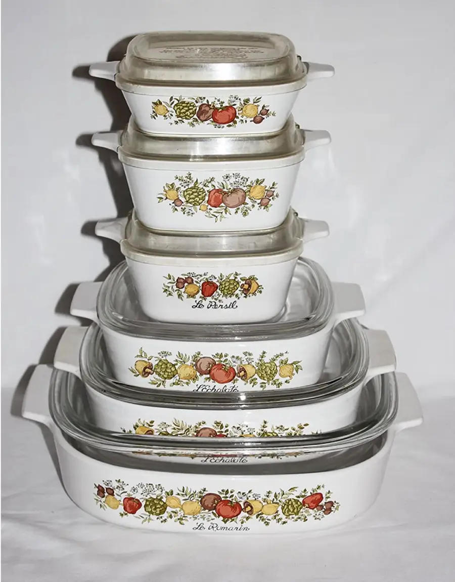 Stack of six vintage Pyrex casserole dishes with lids, each decorated with a colorful vegetable and herb motif labeled “Le Romarin,” on a white background.