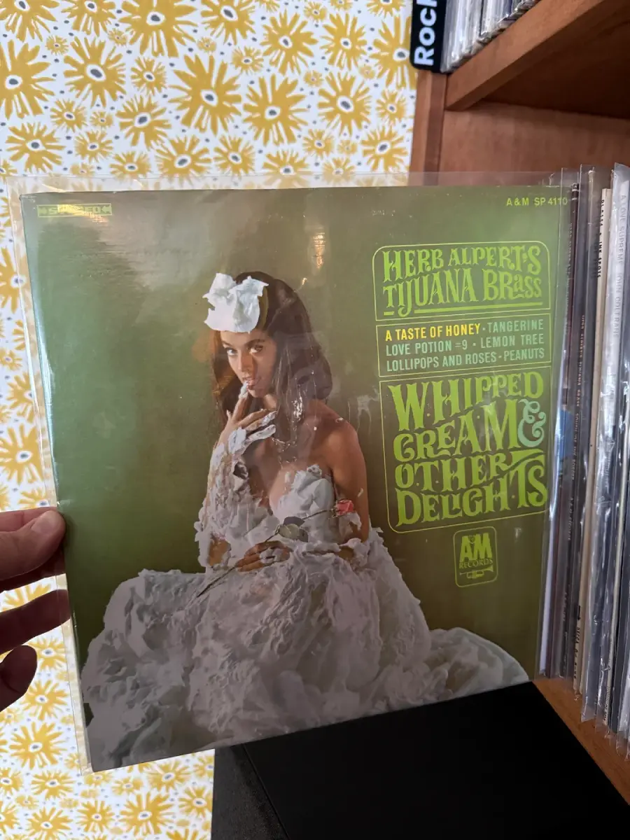 A person in a white dress covered in whipped cream poses on the cover of Herb Alpert's Tijuana Brass album "Whipped Cream & Other Delights." Bold green and yellow text lists songs on the right side.