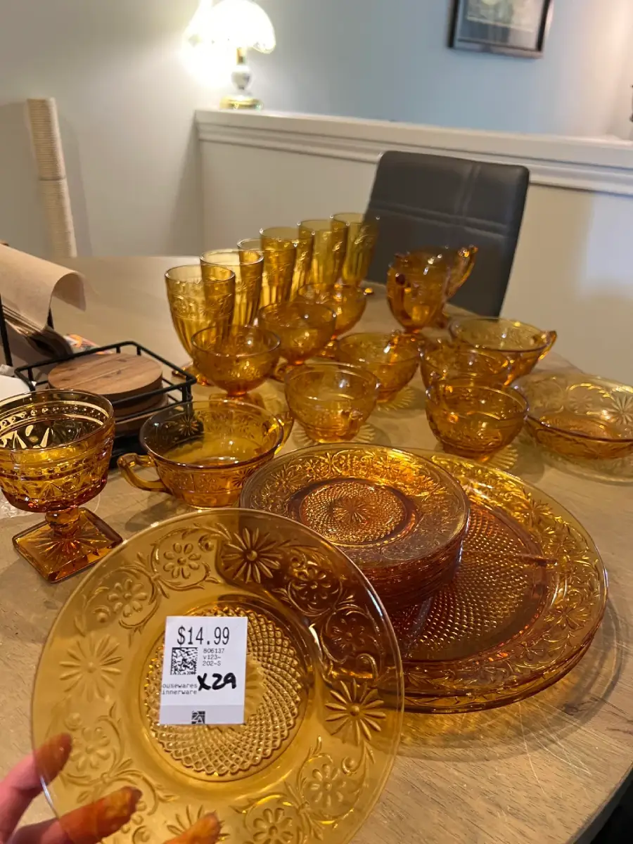 A hand holds an amber glass plate with a $14.99 price tag. More matching vintage amber glassware, including cups and plates, are arranged on a wooden table in a softly lit room.