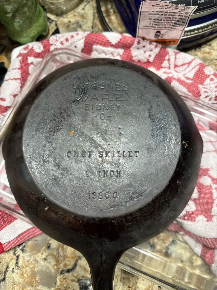 A vintage Wagner Ware Sidney O chef skillet sits upside down on a counter, showing its engraved bottom; text reads "CHEF SKILLET 8 INCH 1380 C." A red and white cloth and other kitchen items are nearby.