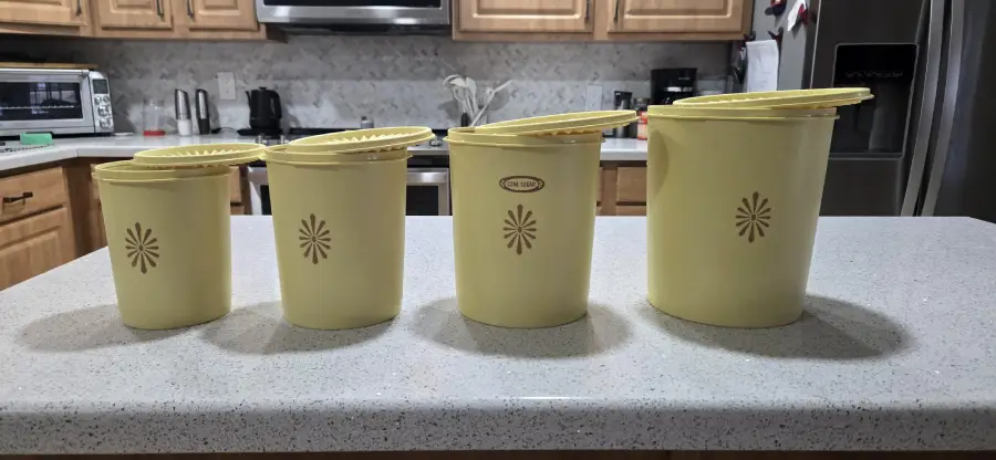 Four yellow Tupperware containers with lids, arranged by size, sit on a white kitchen counter. The kitchen features light wood cabinets, a stove, and a refrigerator in the background.