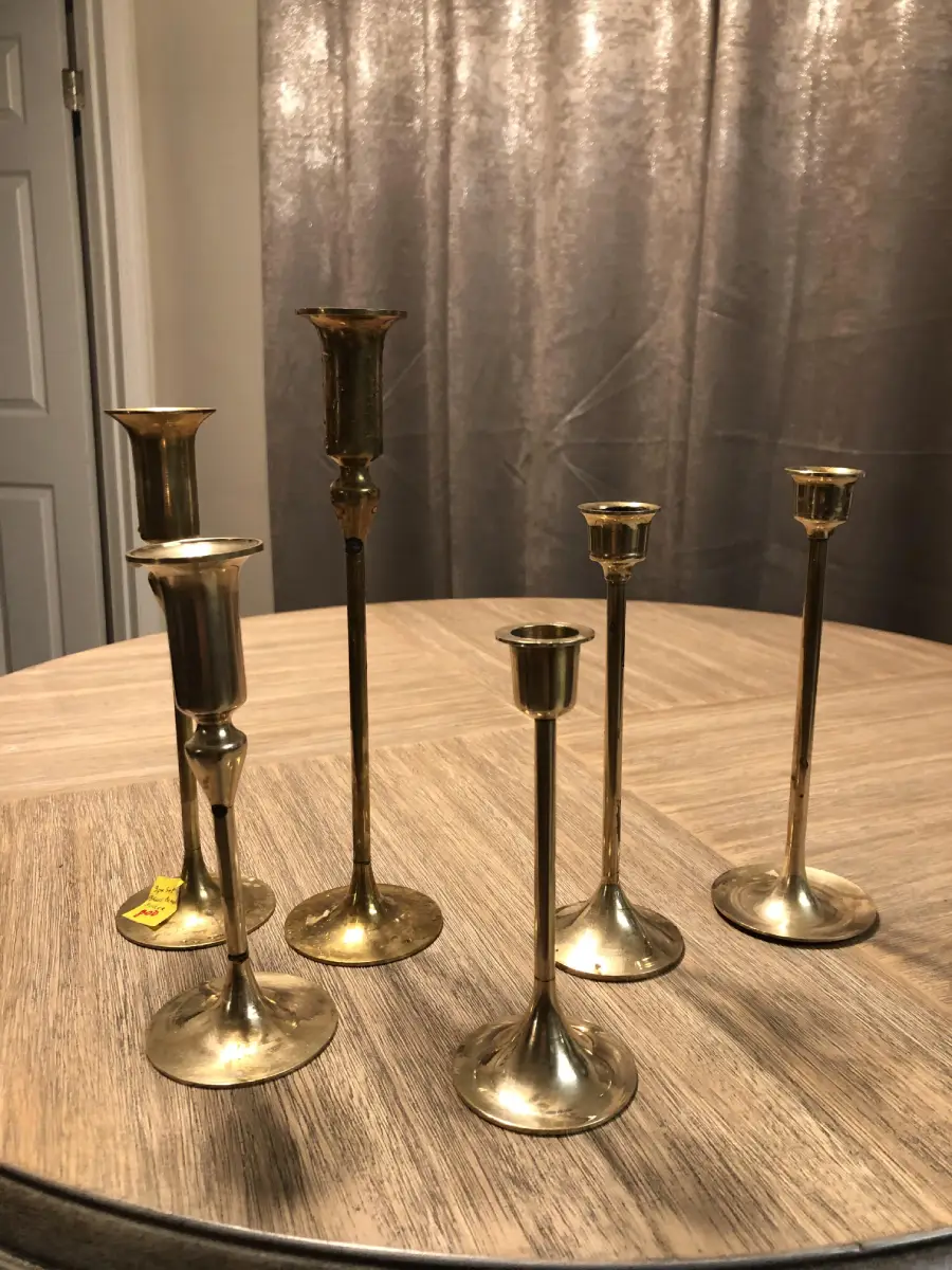 Five brass candlesticks of varying heights are arranged on a light wooden table. A gray curtain and a white door are visible in the background. One candlestick has a yellow price tag.