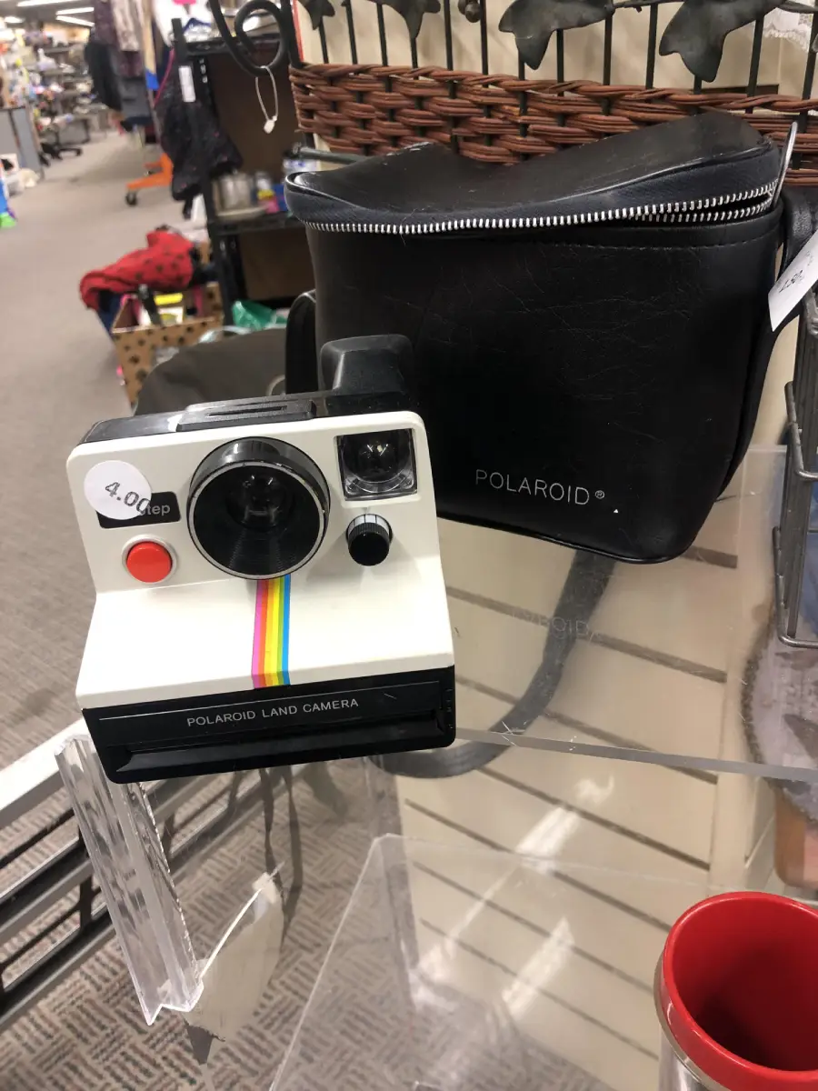 A vintage Polaroid Land Camera with a rainbow stripe sits on display next to a black Polaroid-branded carrying case in a store. A price tag on the camera reads "$9.00".